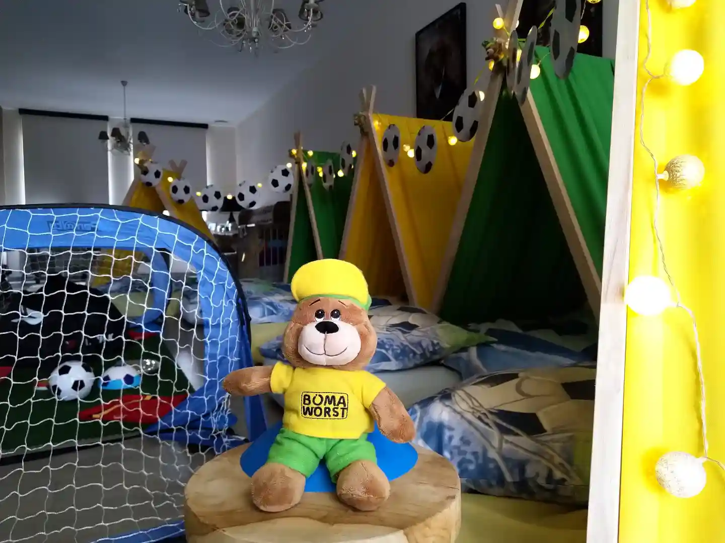 In een woonkamer staan een aantal kleine tentjes in afwisselend geel en groen opgesteld. Voor elk tentje licht een kussen. De nok van de tenten is verbonden met een slinger met voetballen. Vooraan in beeld zit een knuffelbeer in de vorm Jefke, de mascotte van FC De Kampioenen, met een FC De Kampioenen voetbaloutfit aan met op de borst de tekst "Boma Worst". Achter de knuffel staat nog een klein voetbaldoeltje.